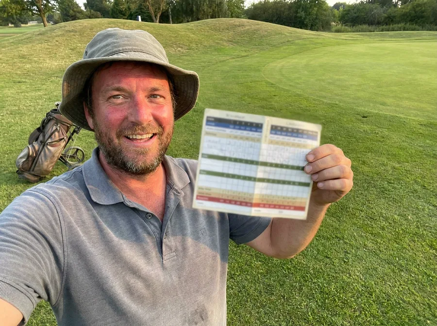 Marcus, in a bucket hat and a very sunburnt grin, holding a scorecard up to the camera on the 18th green.